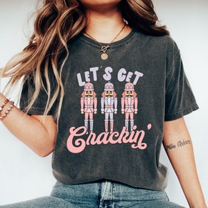 May include: Dark grey t-shirt featuring the phrase "LET'S GET Crackin'" and three nutcracker figures in pink, white, and lavender. A casual style t-shirt suitable for everyday wear.