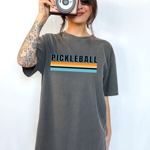 May include: A charcoal grey oversized t-shirt with a retro striped design and the text "Pickleball" in black.