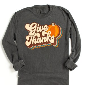 May include: Dark grey long-sleeved shirt featuring the words "Give Thanks" in a retro font, with a pumpkin graphic. The design includes orange, brown, and teal accents, ideal for autumn.