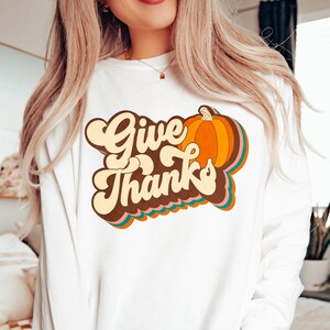May include: White long-sleeved top featuring the words "Give Thanks" in a retro style, alongside a colourful pumpkin illustration. The design incorporates orange, brown, and teal hues. A seasonal autumn-themed garment.