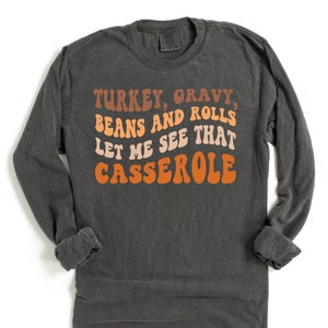 May include: Dark gray long-sleeve shirt with the text "TURKEY, GRAVY, BEANS AND ROLLS LET ME SEE THAT CASSEROLE" in retro orange, brown, and cream colors. The shirt is a casual style.