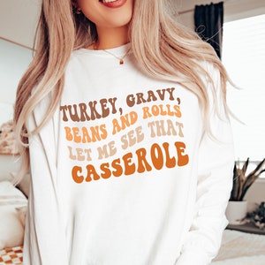 May include: White long-sleeve shirt with the text "TURKEY, GRAVY, BEANS AND ROLLS, LET ME SEE THAT CASSEROLE" in brown, orange, and tan lettering. The shirt is a casual style, suitable for fall or Thanksgiving.