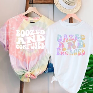 May include: Two tie-dye t-shirts with a white graphic design. The first shirt is pink, yellow, and blue with the text "Boozed and Confused". The second shirt is white with the text "Dazed and Engaged".
