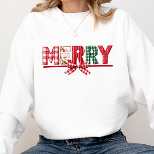 May include: White sweatshirt with the word "MERRY" in red and green plaid letters. A red plaid bow is below the word. The sweatshirt is paired with blue jeans and black sneakers.