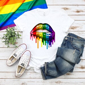 May include: A white t-shirt with a rainbow dripping lips graphic. The shirt is laying on a wooden surface with a pair of white sneakers and blue jeans.