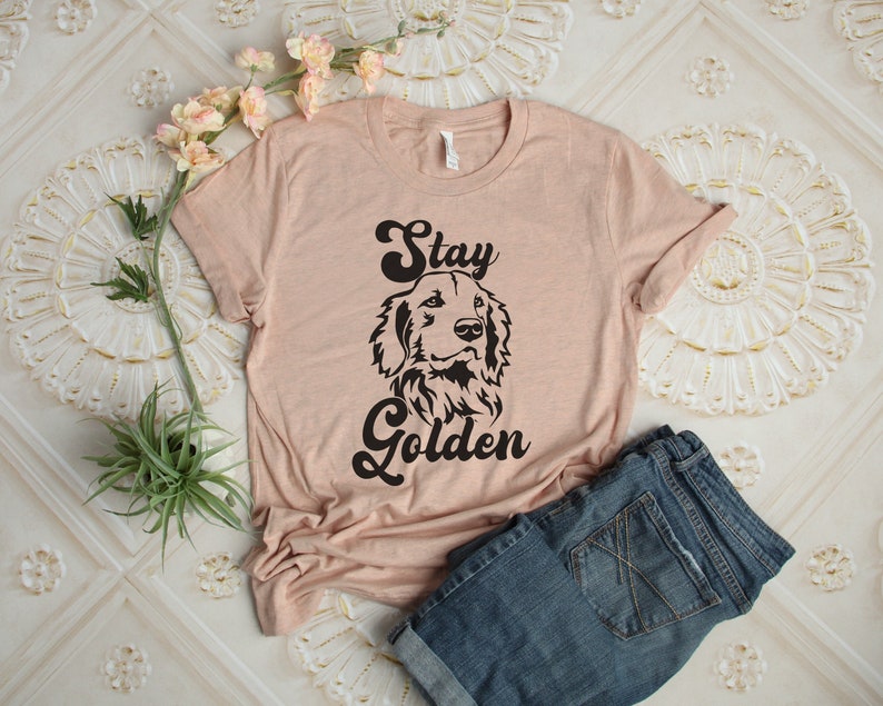 K&ouml;nnte beinhalten: Ein hellrosa T-Shirt mit einer schwarzen Silhouette eines Golden Retrievers und dem Text "Stay Golden".