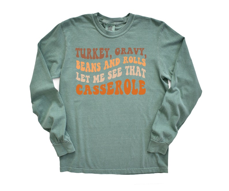 May include: A long-sleeved, sage green shirt with the text "TURKEY, GRAVY, BEANS AND ROLLS, LET ME SEE THAT CASSEROLE" in retro-style orange, brown, and tan lettering. The shirt has a crew neck and long sleeves.
