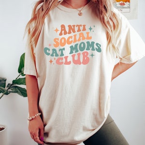Może przedstawiać: Jasnobeżowy T-shirt z napisem "Anti Social Cat Moms Club" w retro czcionce z kolorowymi akcentami. Tekst jest w jasnoniebieskim, pomarańczowym i r&oacute;żowym schemacie kolor&oacute;w.