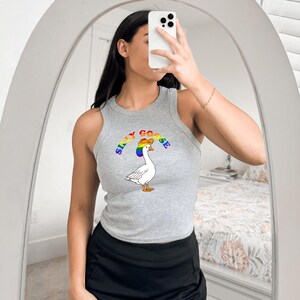 Op de afbeelding: Een grijze tanktop met een cartoon gans met een regenboog hoed en de woorden "Silly Goose" in regenboog letters. De tanktop is mouwloos en heeft een ronde halslijn. De gans is wit met een gele snavel en bruine poten.