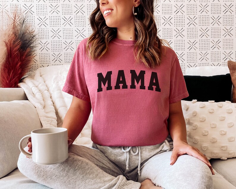 Może przedstawiać: Kobieta w malinowym t-shircie z nadrukiem "MAMA" w kolorze czarnym na przodzie. Siedzi na kanapie z białym kocem i białą poduszką o fakturowanej powierzchni.