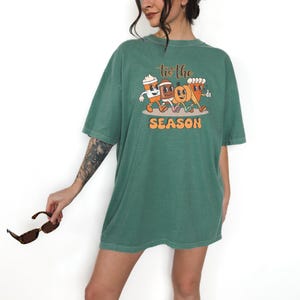 Op de afbeelding: Een groen t-shirt met de tekst "tis the SEASON" en een afbeelding van een koffiekopje, een voetbal, een pompoen en een gebakje. Het shirt heeft een losse pasvorm en korte mouwen.