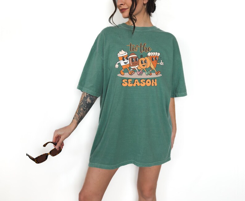 Op de afbeelding: Een groen t-shirt met de tekst "tis the SEASON" en een afbeelding van een koffiekopje, een voetbal, een pompoen en een gebakje. Het shirt heeft een losse pasvorm en korte mouwen.