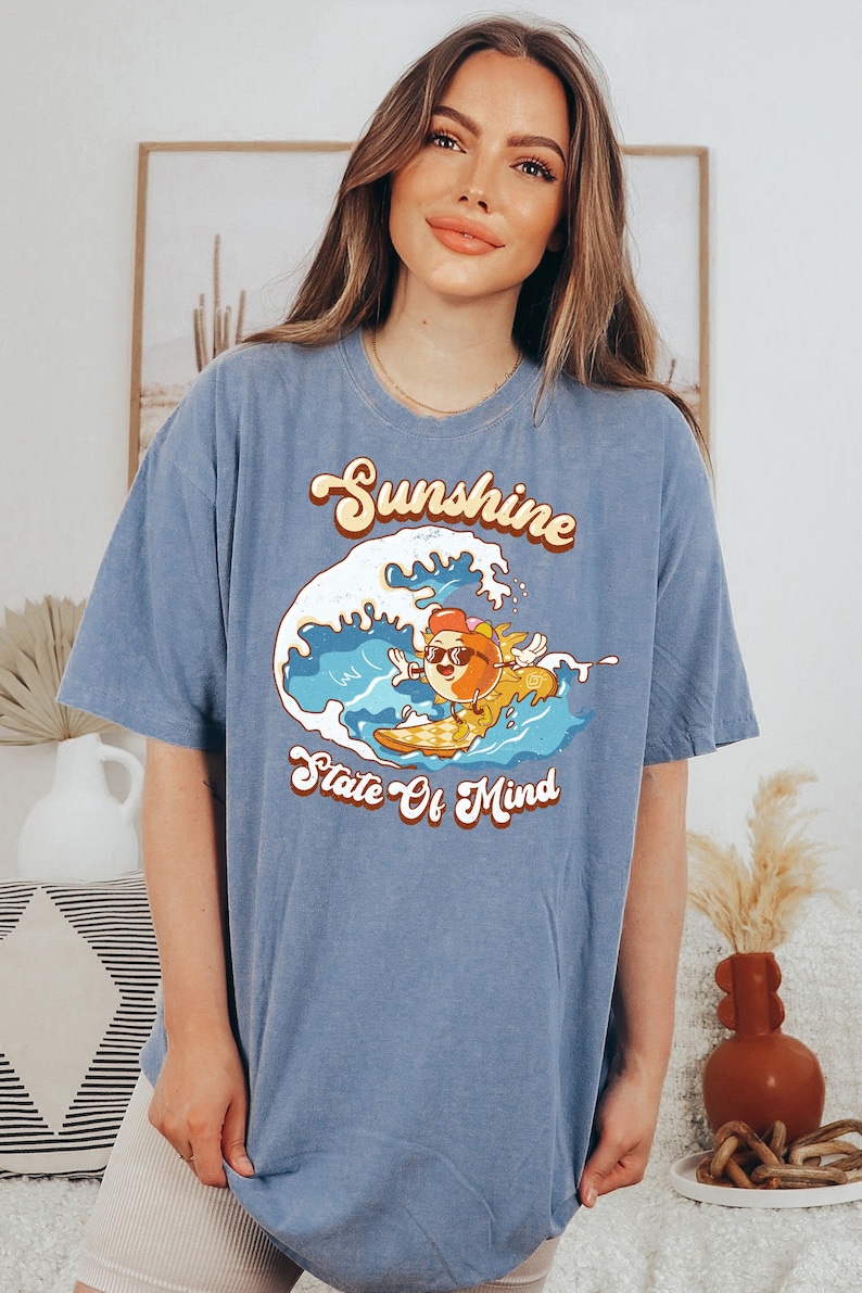 Puede incluir: Una camiseta azul con un gr&aacute;fico de un sol de dibujos animados con gafas de sol y haciendo surf en una ola. El texto "Sunshine State Of Mind" est&aacute; impreso en la camiseta.