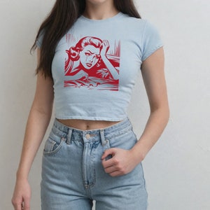 Pu&ograve; includere: Una maglietta crop top blu chiaro con una grafica rossa di una donna che piange. La grafica &egrave; in stile fumetto.