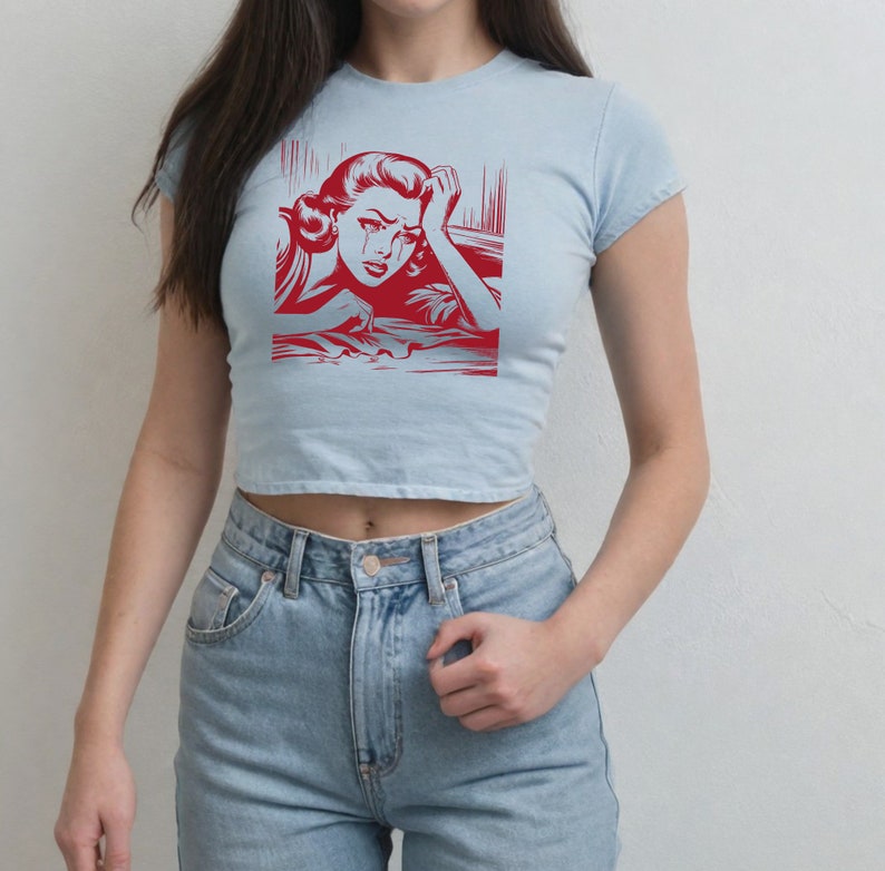 Pu&ograve; includere: Una maglietta crop top blu chiaro con una grafica rossa di una donna che piange. La grafica &egrave; in stile fumetto.