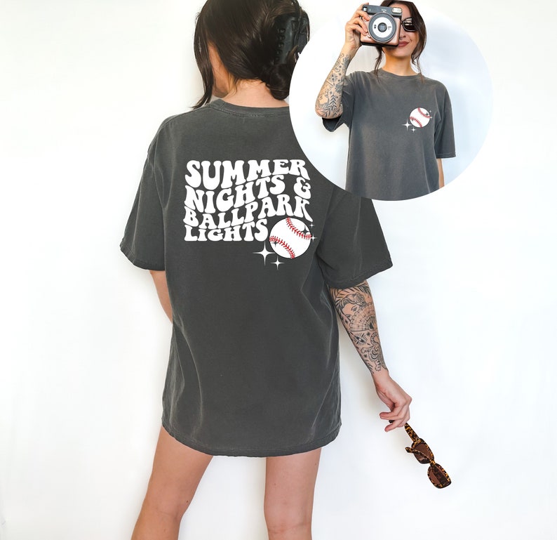 Pu&ograve; includere: Una maglietta oversize di colore grigio scuro con una grafica bianca che dice "Summer Nights & Ballpark Lights" con un grafico di una palla da baseball.