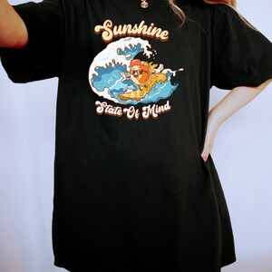 Puede incluir: Camiseta negra de gran tama&ntilde;o con un gr&aacute;fico de un personaje de dibujos animados surfeando en una ola. El texto "Sunshine State Of Mind" est&aacute; impreso en la camiseta.