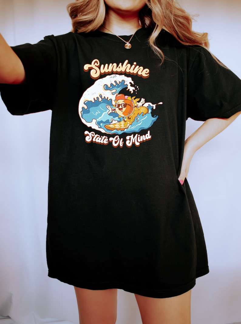 Puede incluir: Camiseta negra de gran tama&ntilde;o con un gr&aacute;fico de un personaje de dibujos animados surfeando en una ola. El texto "Sunshine State Of Mind" est&aacute; impreso en la camiseta.