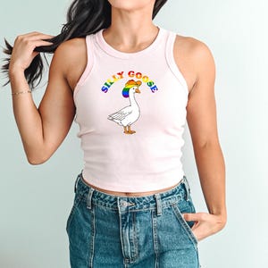 Op de afbeelding: Een lichtroze tanktop met een cartoon gans met een regenboog cowboyhoed. De tekst "SILLY GOOSE" staat boven de gans. De tanktop wordt gecombineerd met een blauwe spijkerbroek.
