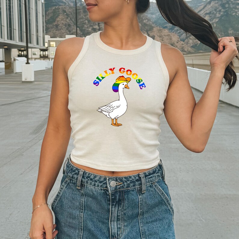 Op de afbeelding: Cr&egrave;mekleurige tanktop met een cartoon gans met een regenboog cowboyhoed. De tekst "SILLY GOOSE" staat in regenboogkleuren boven de gans. De tanktop is mouwloos en heeft een ronde halslijn.
