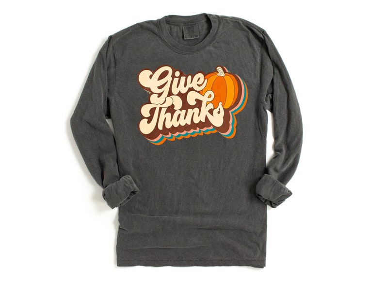 May include: Dark grey long-sleeved shirt featuring the words "Give Thanks" in a retro font, with a pumpkin graphic. The design includes orange, brown, and teal accents, ideal for autumn.