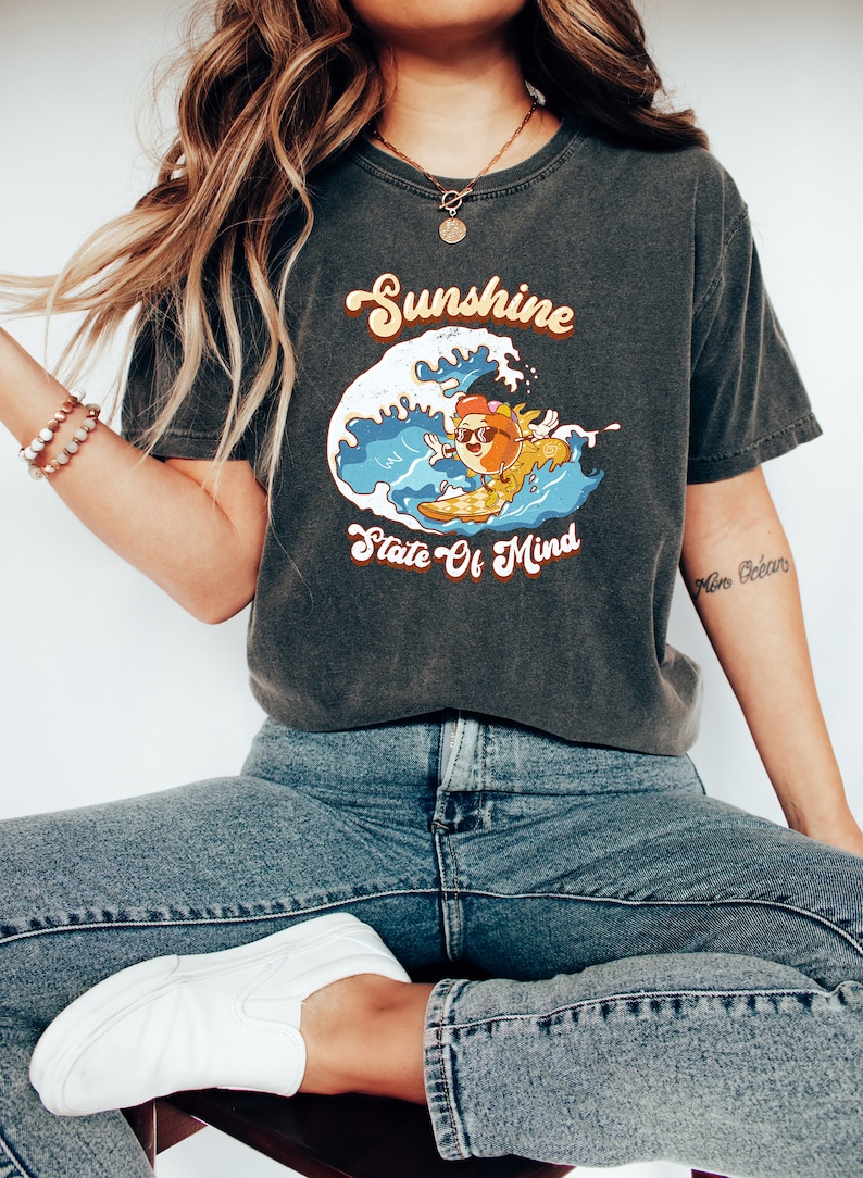 Puede incluir: Una camiseta gris oscuro con un gr&aacute;fico retro de una naranja de dibujos animados con gafas de sol y montando una tabla de surf en una ola. El texto "Sunshine State Of Mind" est&aacute; impreso debajo del gr&aacute;fico.