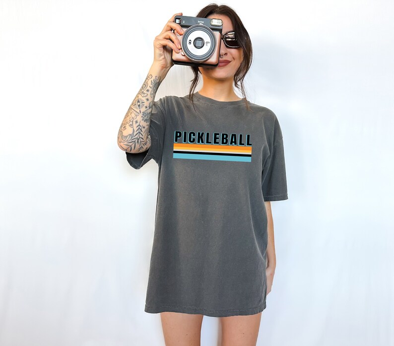 May include: A charcoal grey oversized t-shirt with a retro striped design and the text "Pickleball" in black.