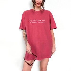 May include: A woman wearing a red oversized t-shirt with the text "I'll just have the chicken tenders" printed on the front.