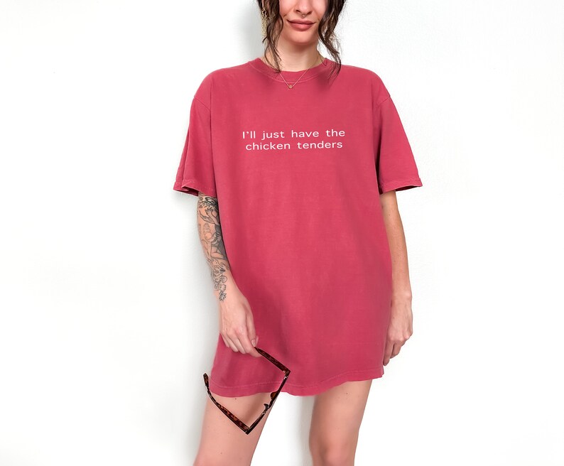 May include: A woman wearing a red oversized t-shirt with the text "I'll just have the chicken tenders" printed on the front.