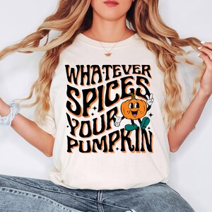 May include: Cream-colored t-shirt with the text "WHATEVER SPICES YOUR PUMPKIN" in black and orange. A cartoon pumpkin with a smiling face and green shoes is in the center. The shirt is a casual, fall-themed design.
