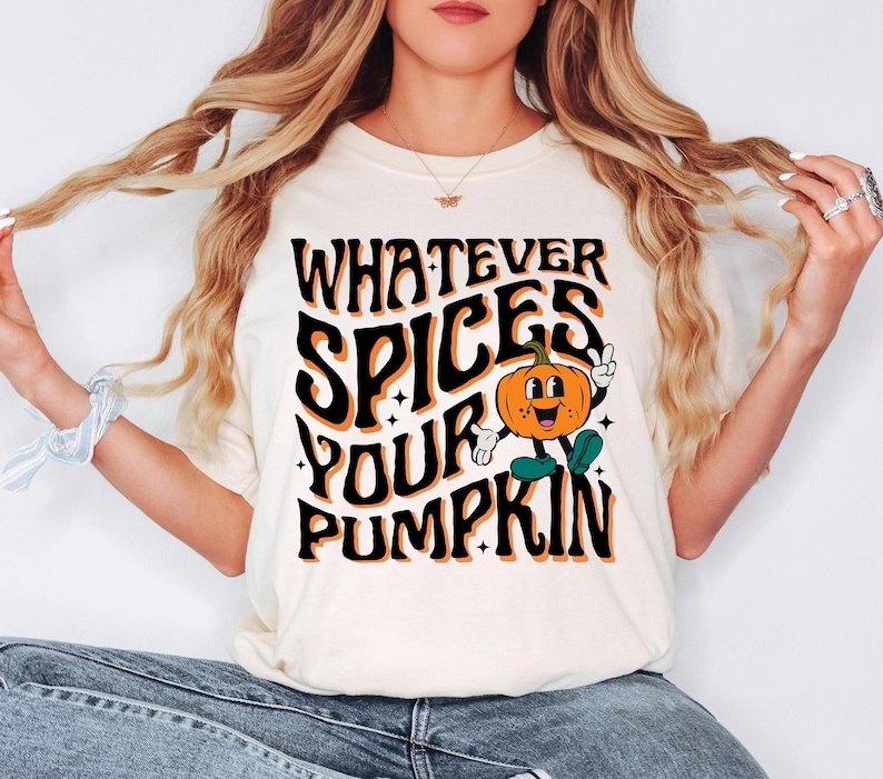 May include: Cream-colored t-shirt with the text "WHATEVER SPICES YOUR PUMPKIN" in black and orange. A cartoon pumpkin with a smiling face and green shoes is in the center. The shirt is a casual, fall-themed design.