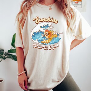 Puede incluir: Una camiseta beige con un gr&aacute;fico de un sol de dibujos animados con gafas de sol y haciendo surf en una ola. El texto "Sunshine State Of Mind" est&aacute; impreso en la camiseta.