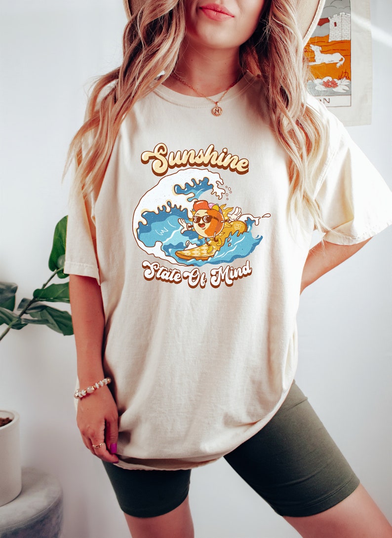 Puede incluir: Una camiseta beige con un gr&aacute;fico de un sol de dibujos animados con gafas de sol y haciendo surf en una ola. El texto "Sunshine State Of Mind" est&aacute; impreso en la camiseta.