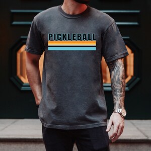 May include: A charcoal grey t-shirt with the word "PICKLEBALL" printed in black with a blue, orange, and black stripe design.