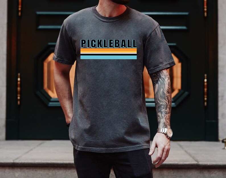 May include: A charcoal grey t-shirt with the word "PICKLEBALL" printed in black with a blue, orange, and black stripe design.