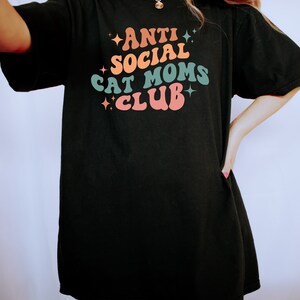 Może przedstawiać: Czarny, oversize'owy T-shirt z napisem "ANTI SOCIAL CAT MOMS CLUB" w kolorowej, retro czcionce.