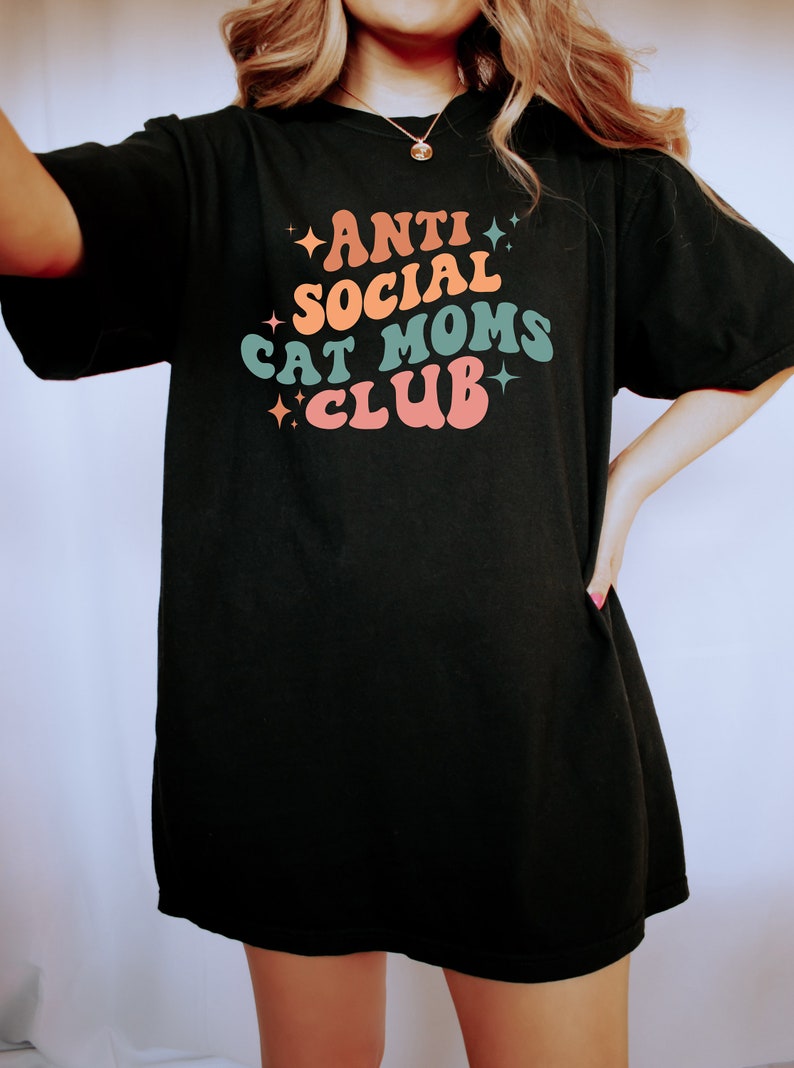 Może przedstawiać: Czarny, oversize'owy T-shirt z napisem "ANTI SOCIAL CAT MOMS CLUB" w kolorowej, retro czcionce.