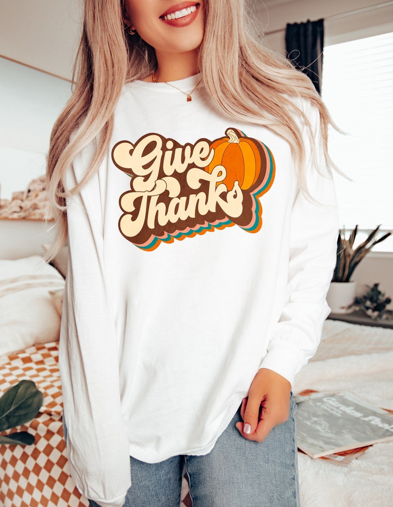 May include: White long-sleeved top featuring the words "Give Thanks" in a retro style, alongside a colourful pumpkin illustration. The design incorporates orange, brown, and teal hues. A seasonal autumn-themed garment.