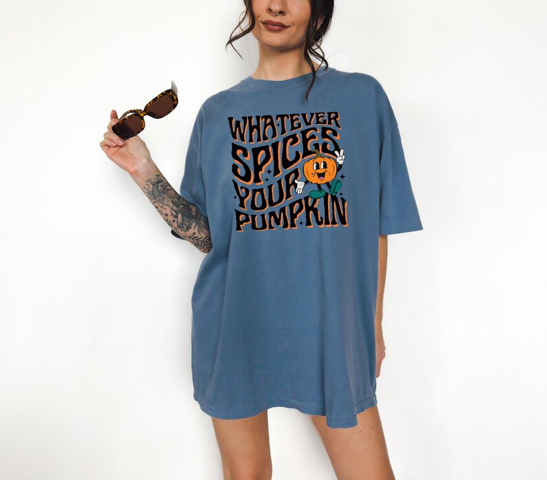 May include: A blue t-shirt with the text "WHATEVER SPICES YOUR PUMPKIN" in black and orange. The graphic includes a smiling pumpkin with green leaves. The shirt is short-sleeved and has a relaxed fit.