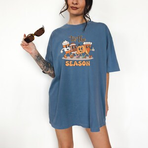 Op de afbeelding: Een blauw t-shirt met een afbeelding van geanimeerde herfstfiguren, waaronder een koffiekopje, een pompoen en een ma&iuml;skolf. De tekst "Tis the Season" staat boven de figuren en "SEASON" eronder. Het t-shirt wordt door een persoon gedragen.