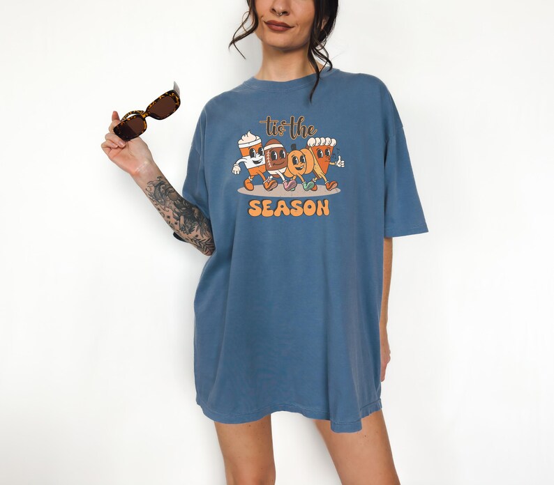 Op de afbeelding: Een blauw t-shirt met een afbeelding van geanimeerde herfstfiguren, waaronder een koffiekopje, een pompoen en een ma&iuml;skolf. De tekst "Tis the Season" staat boven de figuren en "SEASON" eronder. Het t-shirt wordt door een persoon gedragen.