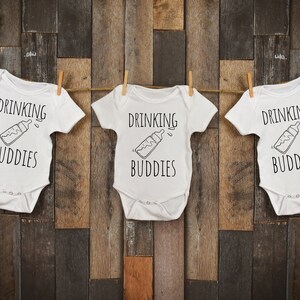 Op de afbeelding: Drie witte baby rompers met de tekst "Drinking Buddies" en een zwart-witte illustratie van een babyfles.