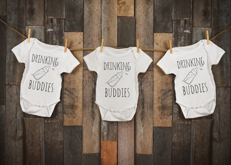 Op de afbeelding: Drie witte baby rompers met de tekst "Drinking Buddies" en een zwart-witte illustratie van een babyfles.