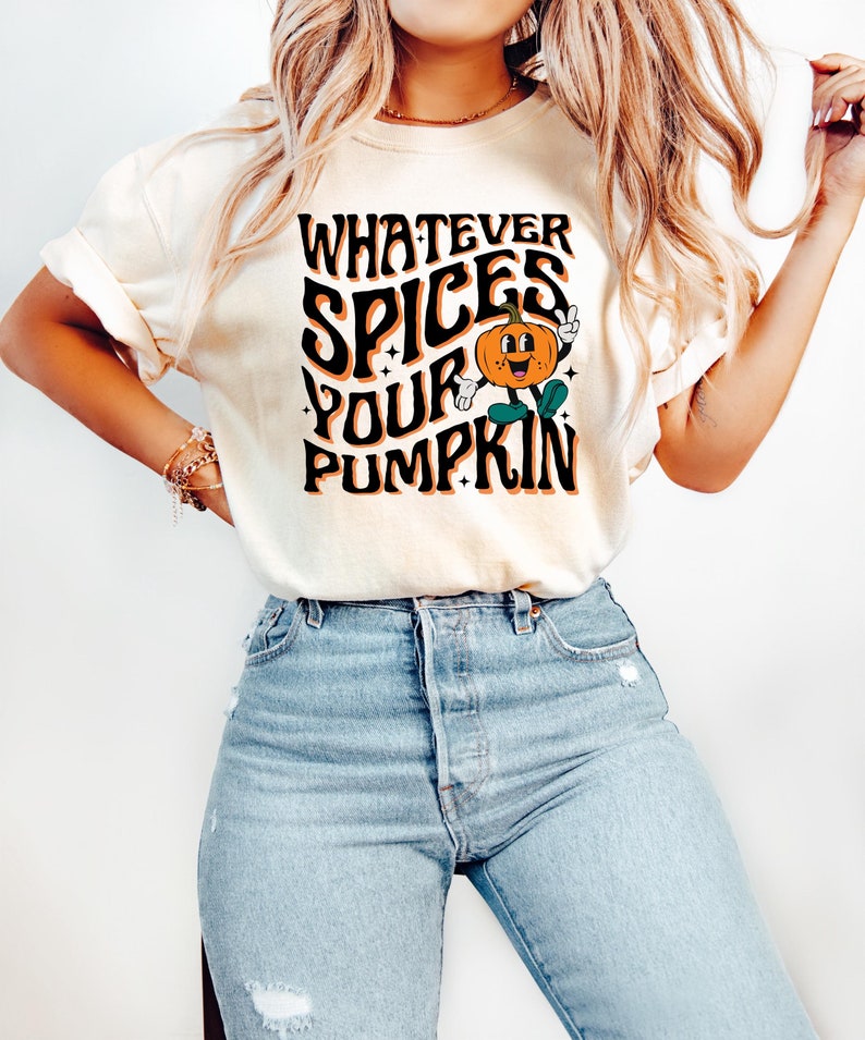 May include: Cream-colored t-shirt with the text "WHATEVER SPICES YOUR PUMPKIN" in black, orange, and white. A cartoon pumpkin character is featured in the design. The shirt is paired with light blue jeans.