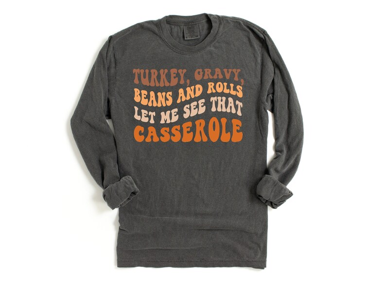 May include: Dark gray long-sleeve shirt with the text "TURKEY, GRAVY, BEANS AND ROLLS LET ME SEE THAT CASSEROLE" in retro orange, brown, and cream colors. The shirt is a casual style.