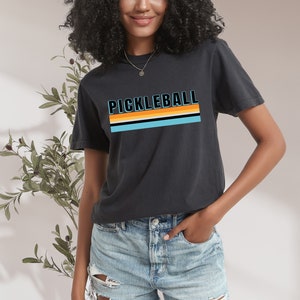 May include: A black t-shirt with a retro-style graphic that says "Pickleball" in blue and orange letters with a striped design underneath.