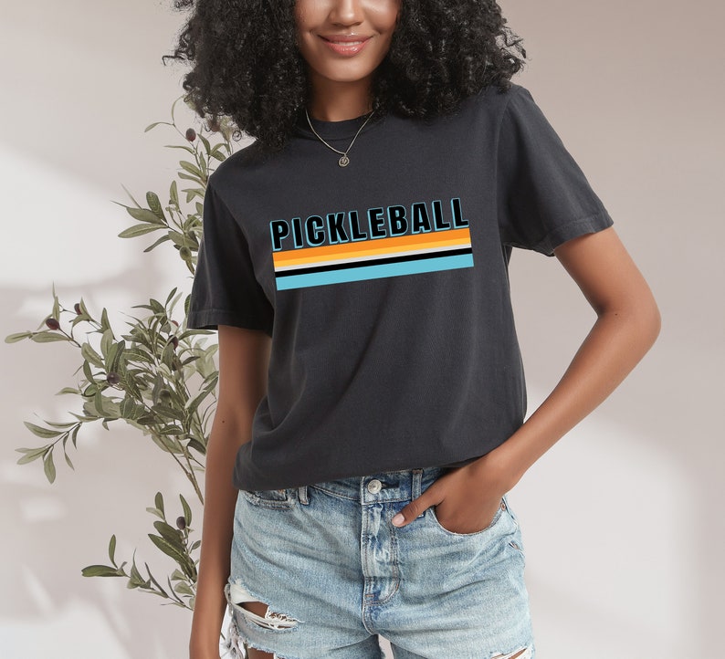 May include: A black t-shirt with a retro-style graphic that says "Pickleball" in blue and orange letters with a striped design underneath.