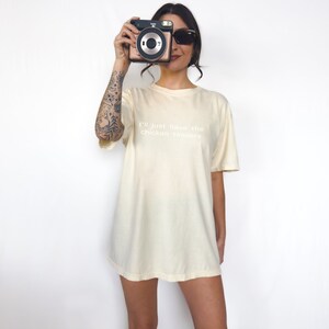 May include: A woman wearing a pale yellow t-shirt with the text "I'll just have the chicken tenders" printed on the front. She is holding a brown and gold instant camera in front of her face.