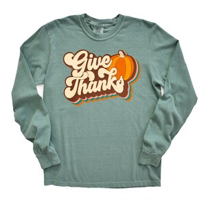 May include: A long-sleeved, teal green shirt featuring the words "Give Thanks" in a retro font, alongside a pumpkin graphic. The pumpkin is orange with brown and orange accents. The shirt is a comfortable, casual style.