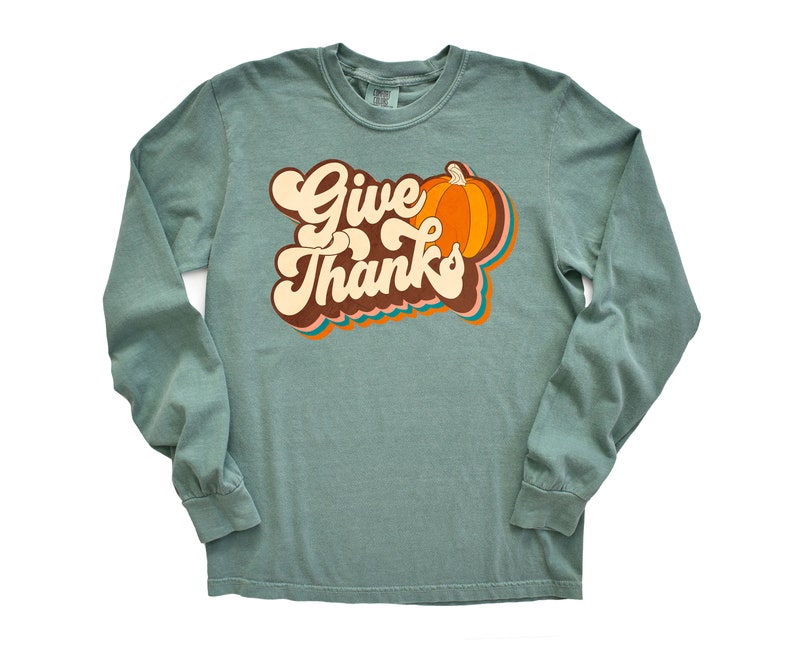 May include: A long-sleeved, teal green shirt featuring the words "Give Thanks" in a retro font, alongside a pumpkin graphic. The pumpkin is orange with brown and orange accents. The shirt is a comfortable, casual style.